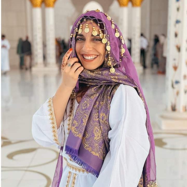 Traveler in a purple and gold coin headscarf smiling in the white marble hall of a grand mosque
